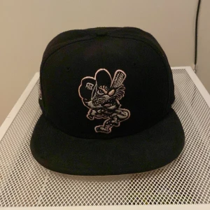 Svart keps med broderad design - Snygg svart keps med en cool broderad design framtill. Kepsen har en platt skärm och en broderad logga på baksidan. Kan gå ner vid snabb affär. 