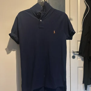 Mörkblå pikétröja från Ralph Lauren - Säljer en stilren mörkblå pikétröja från Ralph Lauren. Tröjan har korta ärmar och en klassisk krage med knappar. Den är tillverkad i bomull och har en liten broderad logotyp på bröstet. Perfekt för en avslappnad men ändå elegant look.