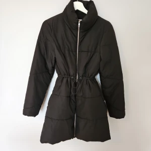 Svart dunjacka från JDY - Snygg svart dunjacka från JDY med dragkedja och justerbar midja. Jackan har en hög krage och långa ärmar, perfekt för kyliga dagar. Enkel och stilren design som passar till det mesta.