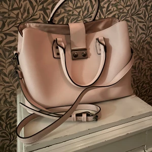 Beige/rosa handväska 👜  - Snygg beige/rosa handväska med justerbar axelrem. Väskan har en stilren design med metallknappar och gott om utrymme för dina saker. Perfekt för en chic och stilren look.