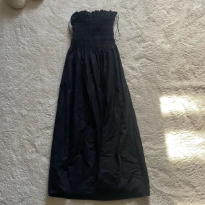 Svart axelbandslös klänning - Snygg svart axelbandslös klänning med smockdetaljer upptill. Perfekt för en stilren look. Klänningen har en lång design och är gjord i ett lätt material som ger en fin passform.