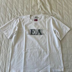 Vit t-shirt Emporio Armani XL - Säljer en stilren vit t-shirt från Armani Exchange med ett tryck av Emporio Armani på framsidan. T-shirten har korta ärmar och är tillverkad i mjukt bomullsmaterial. Perfekt för en avslappnad look. Använd typ 2 gånger, nästan som ny. 