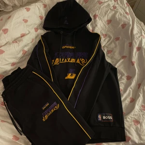 Svart hoodie från BOSS med NBA-tema - Snygg svart hoodie från BOSS med NBA-logga och Lakers-inspirerat tryck i gult och lila. Hoodien har en klassisk design med dragsko i huvan och ribbade muddar. Perfekt för basketfans som vill ha en stilren look.