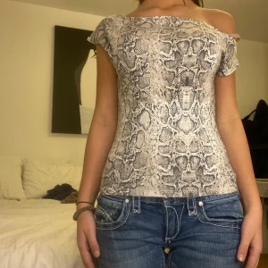 Ormmönstrad offshoulder topp från Nelly - Snygg offshoulder topp med ormmönster i grå och beige toner. Perfekt för en trendig look med jeans eller kjol. Toppen har en tight passform som framhäver figuren. 