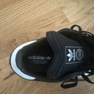 Svarta Adidas Samba sneakers - Snygga svarta Adidas Samba sneakers med klassiska vita ränder och en brun sula. Skorna har snörning och är perfekta för en sportig look. Adidas-loggan syns tydligt på plösen.
