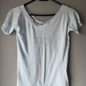 Ljusblå topp från Dolce & Gabbana -  Det står M-L men sitter som s/m.Säljer en ljusblå topp från Dolce & Gabbana med korta ärmar och en v-ringning. Toppen har ett glittrigt DG-märke på framsidan. Perfekt för en stilren look.