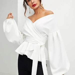 Vit omlottblus med volangdetaljer - Elegant vit omlottblus med offshoulder-design och långa ärmar. Blusen har volangdetaljer och ett knytband i midjan för en smickrande passform. Perfekt för en stilren look. Finns också i svart. 