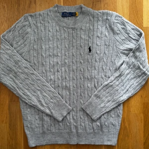 Grå kabelstickad tröja från Ralph Lauren - Säljer en stilren grå kabelstickad tröja från Ralph Lauren. Tröjan har en klassisk design med rund hals och det ikoniska logotypbroderiet på bröstet. Perfekt för en avslappnad men elegant look.