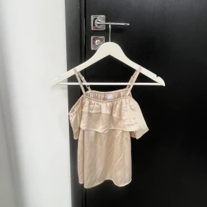 Beige axelbandslös topp - Snygg beige axelbandslös topp med volangdetaljer. Perfekt för varma sommardagar. Tillverkad i ett lätt och luftigt material.