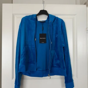 Blå velourjacka från Juicy Couture - Säljer en snygg blå velourjacka från Juicy Couture med dragkedja och huva. Jackan har långa ärmar och en broderad logga på bröstet. Perfekt för en avslappnad stil. 🌀 Helt ny med prislapp. Juicy couture skydiver färgen. Storlek XL. Nypris 1200 kr
