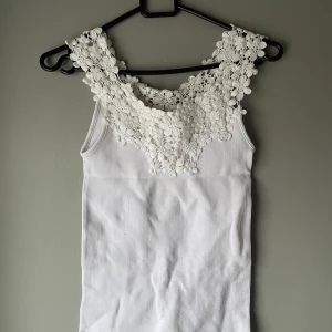 Vit ribbad linnetopp med spetsdetaljer - Säljer en stilren vit ribbad linnetopp med vackra spetsdetaljer vid halsringningen. Perfekt för en somrig look. Toppen är ärmlös och har en elegant design som passar till många tillfällen.