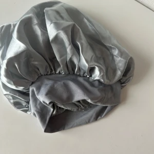 Grå satinmössa/bonett - Elegant grå mössa i satin med en glansig finish. Perfekt för att skydda håret under natten. Den elastiska kanten ger en bekväm passform.