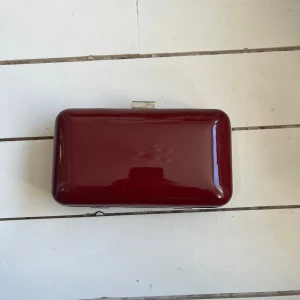 Vinröd kuvertväska - Snygg och stilren vinröd kuvertväska med en glansig yta. Perfekt för att bära med sig det viktigaste. Väskan har en metallknäppning upptill som ger en elegant touch.