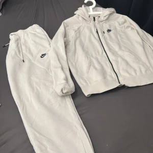 Grå hoodie och byxor från Nike - Säljer en grå hoodie och matchande byxor från Nike. Hoodien har dragkedja och fickor framtill med Nike-logga på bröstet. Byxorna har elastisk midja med dragsko och Nike-logga på benet. Perfekt för en avslappnad stil. Byxa L, Kofts M 