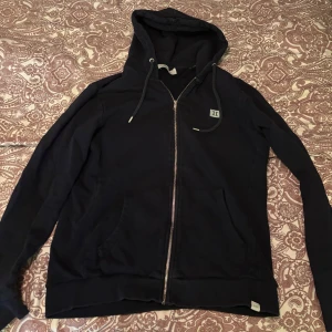 Svart zip från Les Deux - Säljer en stilren svart hoodie från Les Deux med dragkedja och justerbar huva. Perfekt för en avslappnad look. Den har praktiska fickor framtill och ribbade muddar vid ärmslut och nederkant.