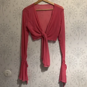 Rosa knytblus - Snygg rosa blus med knyt framtill och långa ärmar. Blusen har volangdetaljer vid ärmsluten och en croppad passform. Perfekt för en trendig look.