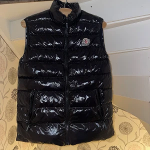 Svart dunväst från Moncler - Snygg svart dunväst från Moncler med glansig yta och dragkedja framtill. Västen har en hög krage och Moncler-logga på bröstet. Perfekt för kyligare dagar när du vill hålla stilen. Den är i mycket bra skick och knappt använd, skriv vid andra frågor eller om du vill ha några specifika bilder 😊 pris kan diskuteras vid snabb och smidig affär 