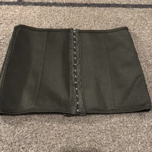 Svart midjekorsett - Säljer en stilren svart midjekorsett med hyskknäppning framtill. Perfekt för att ge en slimmad siluett. Korsetten är enkel och elegant, vilket gör den lätt att matcha med olika outfits.