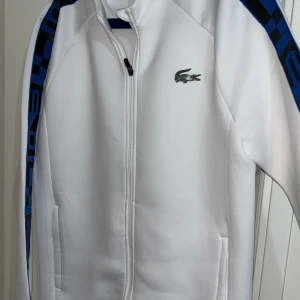 Vit Lacoste tröja  - En vit Lacoste tröja i nylon material. Storleken är Large och modellen av tröjan sitter tajt. Väldigt otydligt fläck (se bild 3), och 2 små slitna delar (se bild 4). Köpt för 1000kr på Lacoste och använd några gånger. 