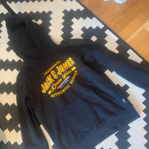 Svart hoodie från Jack & Jones - Säljer en svart hoodie från Jack & Jones med gul text på framsidan. Tröjan har en klassisk känguruficka och en bekväm huva. Perfekt för en avslappnad stil.