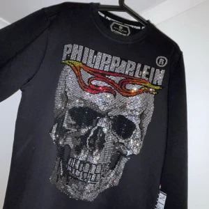 Svart långärmad tröja från Philipp Plein - Snygg svart långärmad tröja från Philipp Plein med en stor glittrig dödskalle och flammor på framsidan. Baksidan har en lista med städer och Philipp Plein-logga i färgglada bokstäver. Perfekt för dig som vill sticka ut!