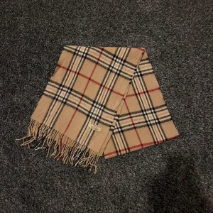 Beige rutig halsduk från Burberry - Snygg beige halsduk från Burberry med klassiskt rutigt mönster i svart, rött och vitt. Tillverkad i 100% lammull för en mjuk och lyxig känsla. Perfekt för att hålla dig varm och stilren under kyliga dagar. (tre jättelitet hål finns men märks inte)