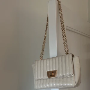 Vit quiltad axelväska med kedja - Snygg vit axelväska med quiltad design och guldfärgad kedja. Väskan har en elegant låsdetalj framtill och är perfekt för att ge en stilren touch till din outfit. Tillverkad i syntetmaterial.