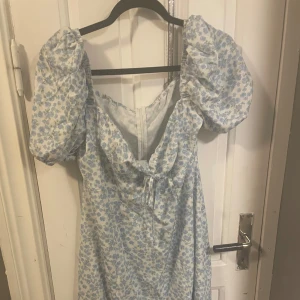 Blommig långklänning med puffärmar - Charmig långklänning med blått blommönster på vit bakgrund. Klänningen har puffiga kortärmar och en söt knytning framtill. Perfekt för sommarens alla tillfällen!