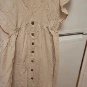 Beige klänning med knappar - Charmig beige klänning med v-ringning och knappar framtill. Den har en luftig passform och korta ärmar, perfekt för varmare dagar. Klänningen har en avslappnad stil med en lätt rynkad design under bysten.