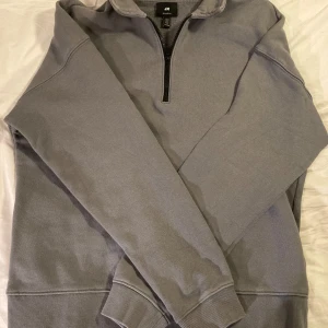 Grå tröja med dragkedja från H&M - Säljer en grå tröja från H&M med halv dragkedja och relaxed fit. Tröjan har långa ärmar och ribbade muddar vid ärmslut och nederkant. Perfekt för en avslappnad stil.