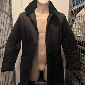 Svart regnrock från H&M - Säljer en stilren svart regnrock från H&M. Rocken har långa ärmar och knappar framtill. Perfekt för höst- och vårsäsongen. Den har en klassisk design med en hög krage och är tillverkad i ett syntetmaterial.