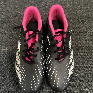 Adidas Predator fotbollsskor - Snygga Adidas Predator fotbollsskor i svart med vita och rosa detaljer. Skorna har snörning och en mönstrad yta för bättre grepp på planen. Perfekta för fotbollsspelare som vill ha stil och funktion.