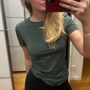 Grön t-shirt från Nike - Snygg grön t-shirt från Nike med korta ärmar och en liten vit logga på bröstet. Perfekt för en sportig look eller träning. T-shirten har en normal passform och är tillverkad i ett mjukt material.