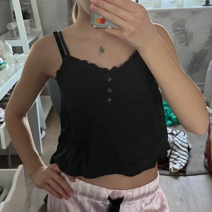 Svart linne från H&M - Säljer en svart linne från H&M med smala axelband och knappar framtill. Toppen har en ribbad struktur och spetsdetaljer vid axelbanden. Färgen är typ mörk grå och lite mörk grön. Perfekt för en sommar dag. Kostade 150 kr när jag köpte den. Använd 1-2 gånger. Super duper söt topp!! 😍😍