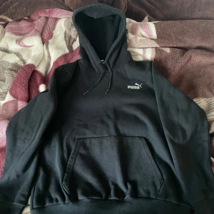 Svart hoodie från Puma - Säljer en klassisk svart hoodie från Puma med en känguruficka framtill och justerbar huva. Perfekt för en avslappnad stil. Puma-loggan är tryckt på bröstet. Passar bra till både vardag och träning.