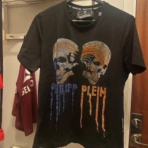 Svart t-shirt med dödskallemotiv från Philipp Plein - Snygg svart t-shirt från Philipp Plein med ett coolt dödskallemotiv i blått och orange på framsidan. T-shirten har korta ärmar och en normal passform. 
