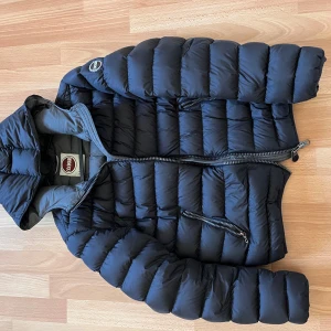 Svart pufferjacka från Colmar - Snygg svart pufferjacka från Colmar med dragkedja och huva. Perfekt för kyliga dagar med sin quiltade design. Jackan har två praktiska fickor med dragkedja framtill. En stilren och funktionell jacka för vintern.