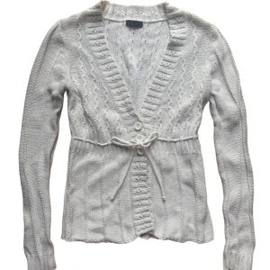 Vit vintage stickad kofta från Vero Moda - Säljer en stilren vit stickad kofta från gammal Vero Moda. Koftan har långa ärmar och fina detaljer med knappar och knytband framtill. Passar en XS-M beroende på önskad passform. 