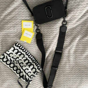 Svart axelväska från Marc Jacobs - Snygg svart axelväska från Marc Jacobs(snapshot crossbody bag)  med justerbar rem och stilren design. Väskan har en praktisk storlek och är perfekt för att bära med sig det viktigaste. Den kommer med originalförpackning och etiketter.