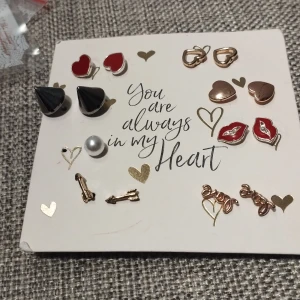 Set med örhängen i olika former - Ett charmigt set med örhängen i olika former och färger. Innehåller hjärtan i rött och guld, svarta konformade örhängen, pärlor, röda läppar och små pilar. Perfekt för att mixa och matcha efter humör och stil.