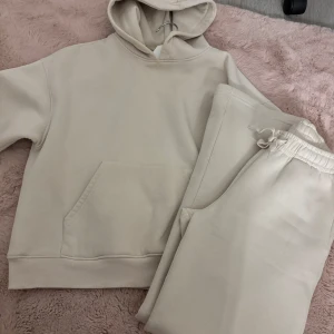 Beige hoodie och byxor set - Säljer ett mysigt set med en beige hoodie och matchande byxor. Hoodien har en klassisk design med känguruficka och ribbade muddar. Byxorna har en bekväm resårmidja med dragsko. Perfekt för en avslappnad stil. Hoodie är i strl xs och byxorna i xxs. Nyskick!