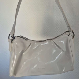 Vit axelväska med dragkedja - Snygg vit axelväska i syntetmaterial med en praktisk dragkedja upptill. Väskan har en stilren design och en bekväm axelrem. Perfekt för att bära dina nödvändigheter med stil.