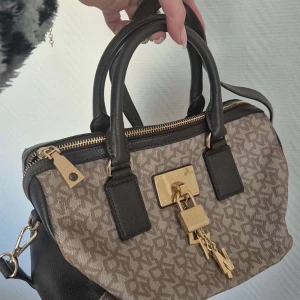 Beige och svart handväska med gulddetaljer - Snygg handväska i beige och svart med gulddetaljer. Väskan har ett mönster och en dekorativ låsdetalj framtill. Den har både handtag och en avtagbar axelrem för flexibilitet. Perfekt för att ge din outfit en lyxig touch.