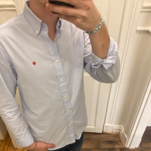 Ljusblå skjorta från Morris - Säljer en stilren ljusblå skjorta från Morris med broderad röd detalj på bröstet. Skjortan har en klassisk krage och knappar framtill. Modellen är ca 182, tveka inte att kontakta vid eventuella frågor 😀