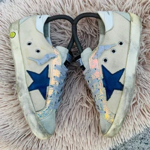 Beige sneakers med blå stjärna - Snygga beige sneakers med en blå stjärna på sidan och glittriga detaljer. Skorna har snörning och en smiley-detalj på hälen. Perfekta för en cool och avslappnad stil.