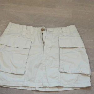 Beige kortkjol med fickor - Säljer en beige kortkjol från lager 157  i storlek XS. Kjolen har stora praktiska fickor framtill och stängs med dragkedja och knapp. Perfekt för en avslappnad stil.