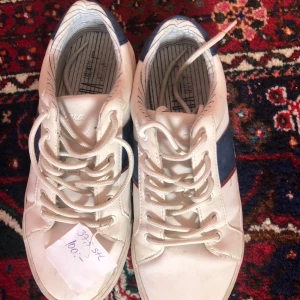 Vita sneakers med blå och röda detaljer - Snygga vita sneakers med snörning och blå samt röda ränder på sidan. Perfekta för en avslappnad stil. Storlek 37.