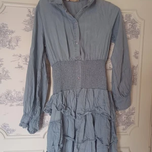 Blå klänning från Pernilla Wahlgren - Charmig blå klänning från Pernilla Wahlgren med långa ärmar och knappar framtill. Den har en smockad midja och volangdetaljer på kjolen som ger en romantisk touch. Perfekt för en stilfull look.