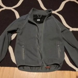 Grågrön fleecejacka från Kuling - Säljer en grågrön fleecejacka från Kuling med dragkedja framtill och två praktiska fickor med dragkedjor. Jackan har långa ärmar och en hög krage för extra värme. Perfekt för kyliga dagar!