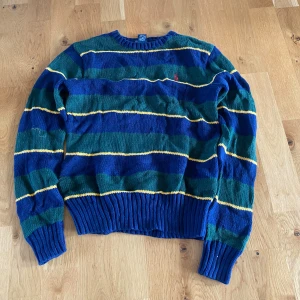 Randig tröja från Polo Ralph Lauren - Säljer en snygg randig tröja från Polo Ralph Lauren i blått, grönt och gult. Tröjan är tillverkad i 100% bomull och har långa ärmar med ribbade muddar. Den har en broderad logotyp på bröstet. Perfekt för en stilren look!